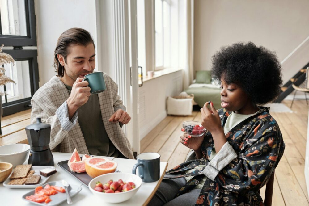 Two people sitting by the table drinking from a cup and eating fruits