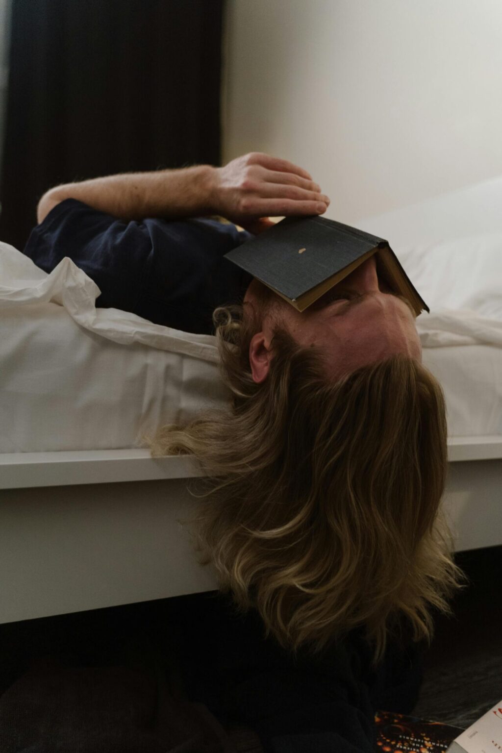 Man lying with head hanging off bed and book on his face