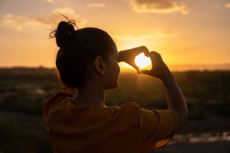 Woman doing hand heart sign in front of the sun