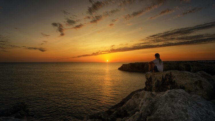 Person sitting on rock next to body of water during sunset