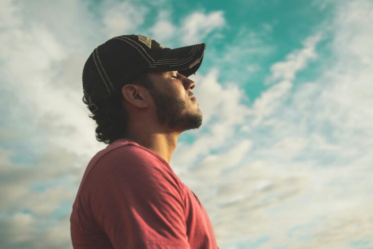 Man wearing black cap with eyes closed under cloudy sky