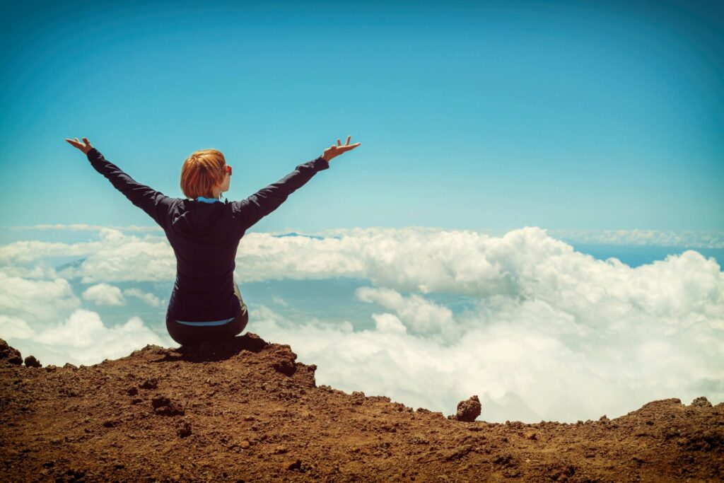 Person sitting on a cliff overseeing clouds and raising up both hands