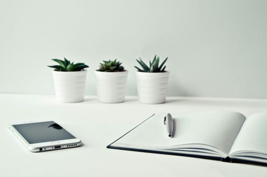 Three white ceramic pots with green leaf plants near open notebook on white table