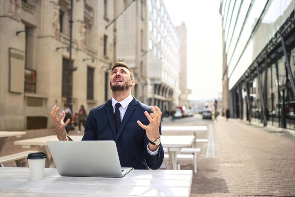 Frustrated man sitting in front of laptop and coffee cup and looking up at the sky