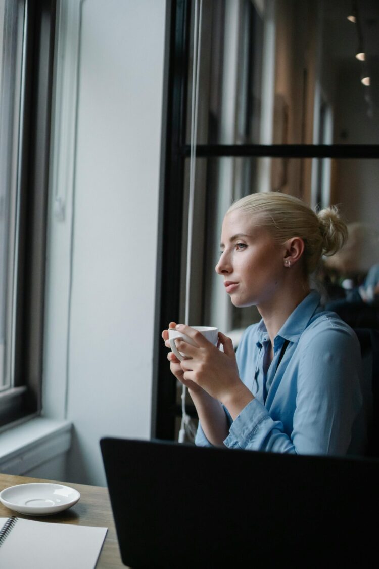 Serious woman drinking coffee in office and looking at window