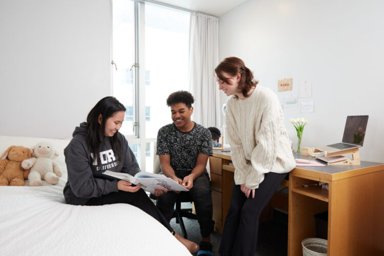 Three students sitting on the bed and chair inside a dorm room.