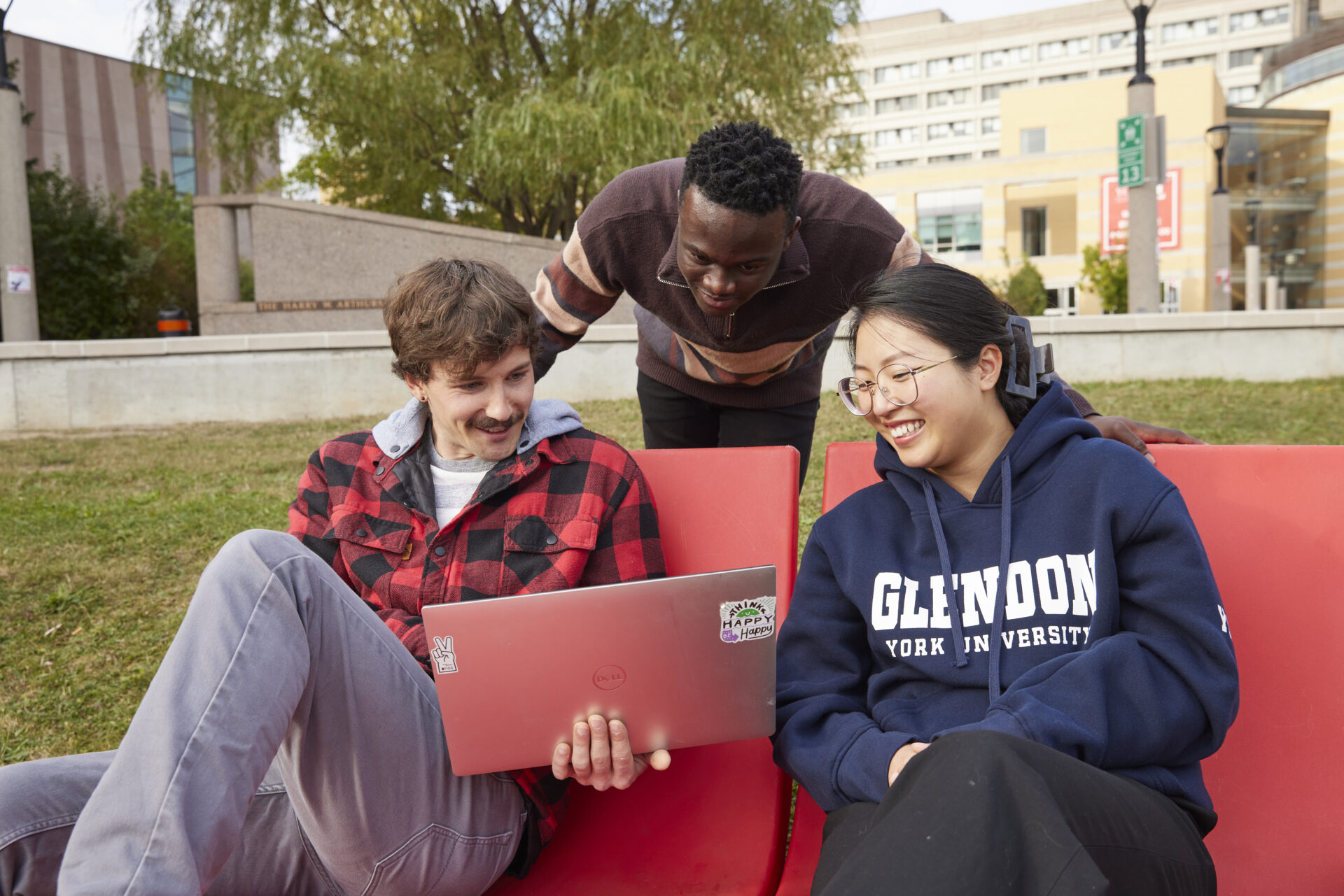 Three students sitting outside on red chairs, looking at a laptop together.