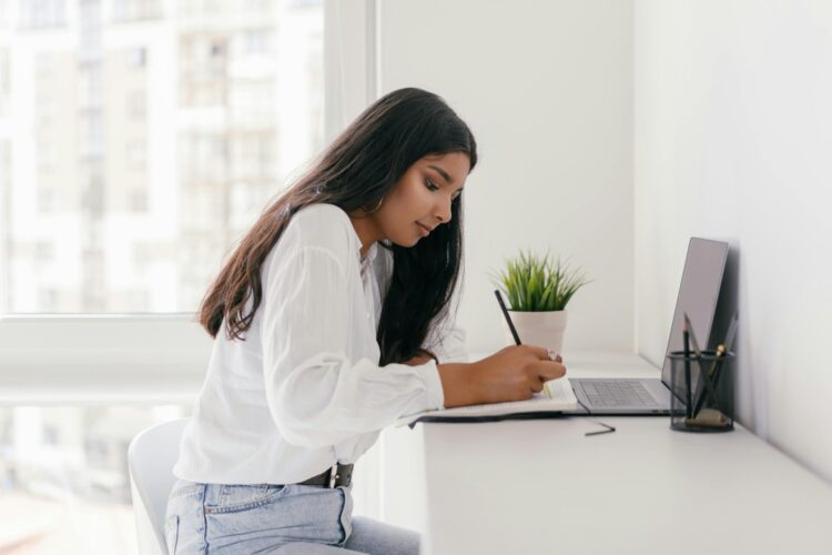 Student writing in a notebook while working on a laptop at a desk.