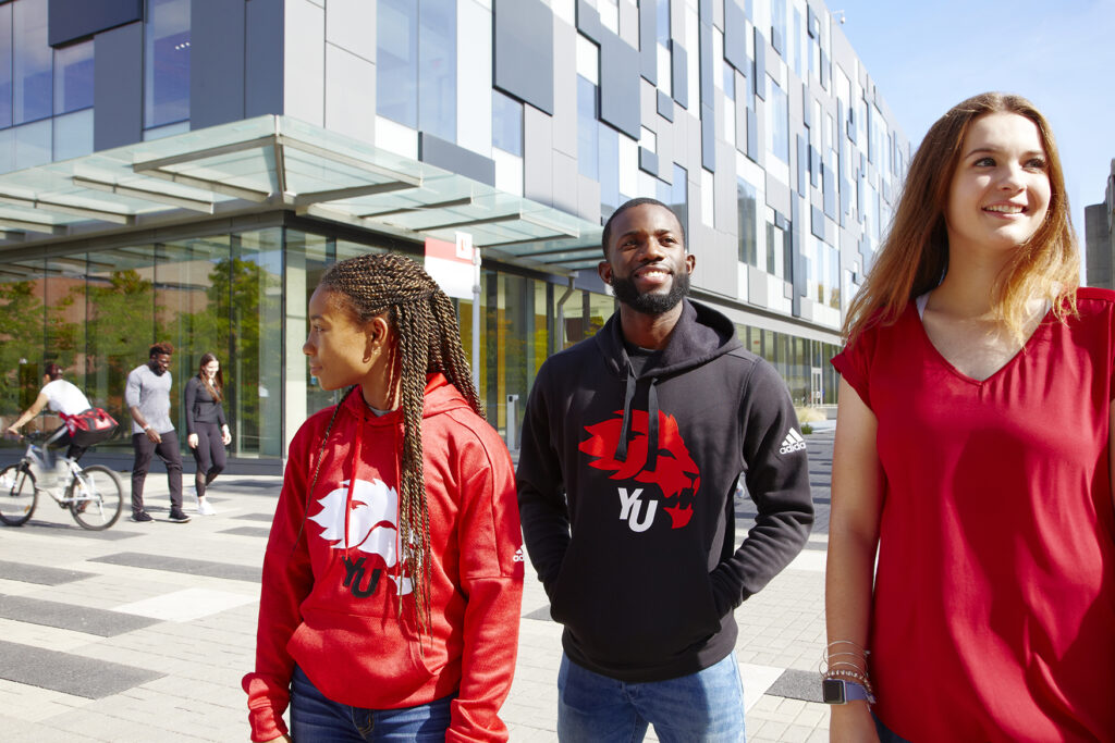 Three students wearing York University apparel walk together outside a glass-front campus building on a sunny day at York University.