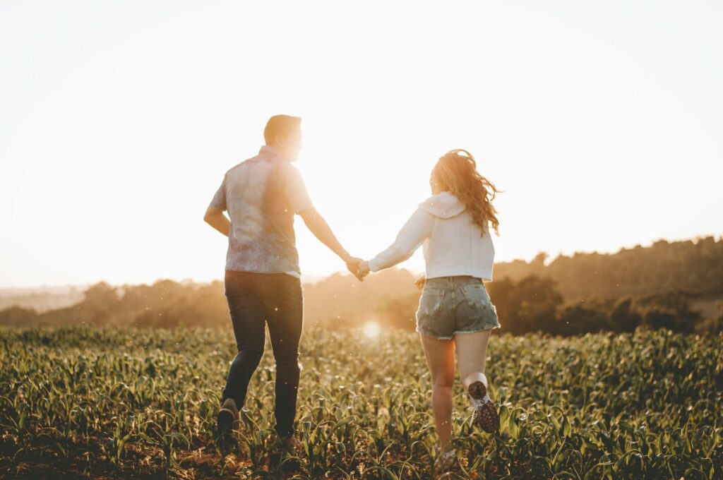 A couple joyfully runs hand in hand through a sunlit field at sunset, capturing a sense of love, freedom, and playful connection in a natural outdoor setting.